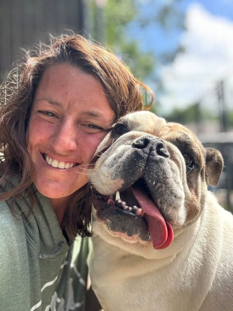 A smiling woman in a green hoodie leans her head against a happy, panting English Bulldog during an outdoor bonding moment.